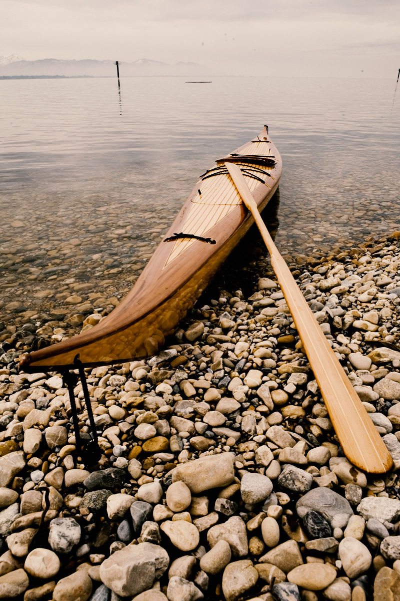 Frontansicht des Ninook-Kajaks am Ufer des Bodensees, mit schräg nach vorne gerichtetem Paddel und Bergen im Hintergrund.